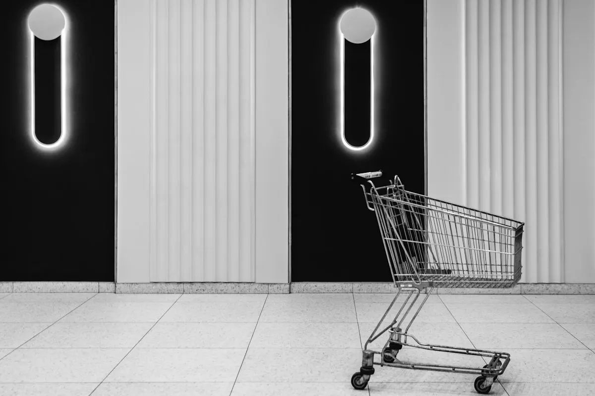 A shopping cart is placed on a tiled floor in front of a modern wall with vertical lines and glowing circular lights resembling exclamation marks. The overall setting is minimalist with a monochrome color scheme.
